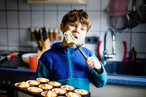 Cute little happy blond school kid boy baking carrot blueberry muffins in domestic kitchen, indoors. Funny lovely healthy child having fun, tasting sweet dough. Little helper.