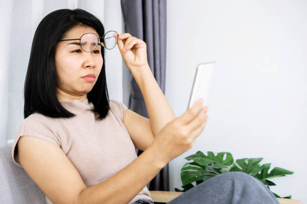 mujer asiática sosteniendo los anteojos teniendo problemas con el visionado tratando de leer texto en el teléfono inteligente - entrecerrar los ojos fotografías e imágenes de stock