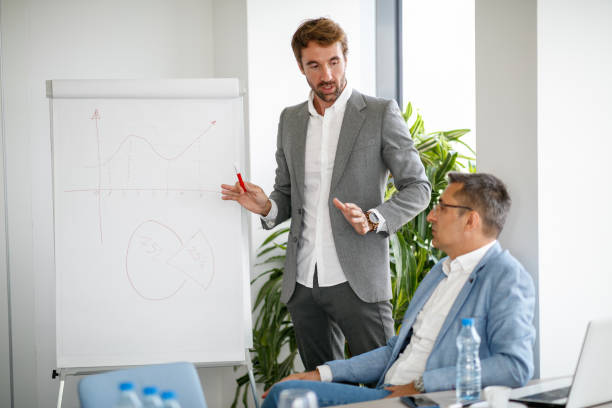 istock Young man by the flipchart explaining situation shown on graph to his colleague 1307919491