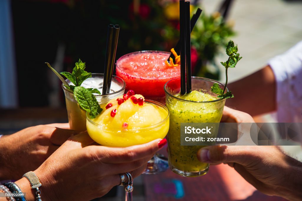 Four hands holding glasses with yellow and red fruit cocktails in a toast Four hands holding drinking glasses with yellow and red fruit cocktails, proposing a toast at outdoor party Cocktail Stock Photo