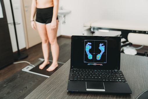 Young adult woman is standing on a medical pressure scanner to analyze her footprint and realize new shoe insoles to improve her posture. She's in a medical clinic. A computer with feet's graphs in the foreground.