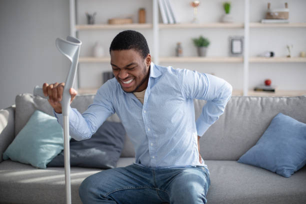 istock Young black guy suffering from lower back pain, using crutch, sitting on sofa at home. Healthcare and medical concept 1307929562