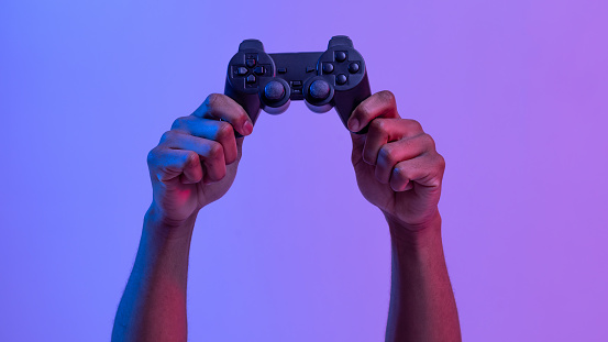 Black male hands holding joystick for video games over purple background with neon lighting, unrecognizable man enjoying modern technologies for gaming, cropped image, panorama with copy space