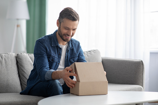 Happy middle-aged man unpacking delivery box, sitting on couch by tea table, home interior. Satisfied customer cheerful man opening package, ordering clothes or gadgets on Internet, copy space