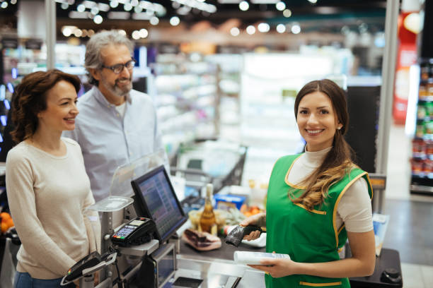 istock Young cashier checking out customers at supermarket while facing camera smiling 1307933281