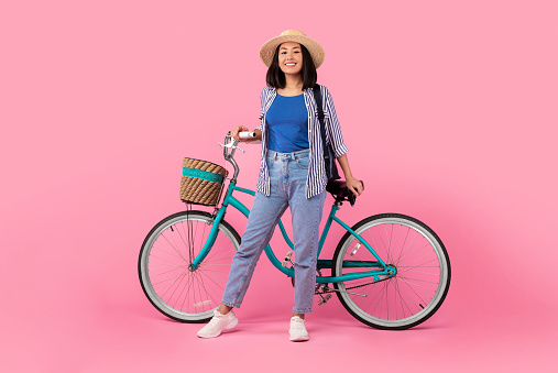 Springtime Activity. Full body length shot of trendy asian lady wearing summer hat and backpack posing and holding handlebar of classic bicycle with wicker basket isolated on pink studio background