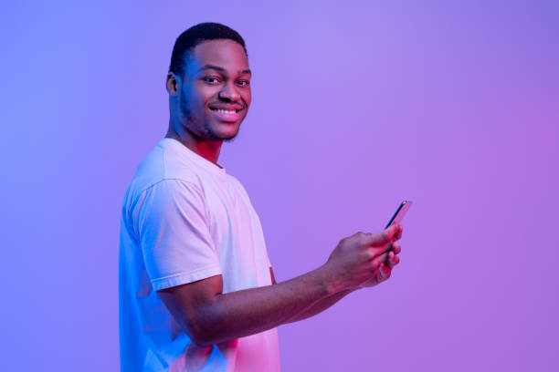 istock Happy Black Man Using Smartphone, Standing Under Neon Light Over Purple Background 1307934969