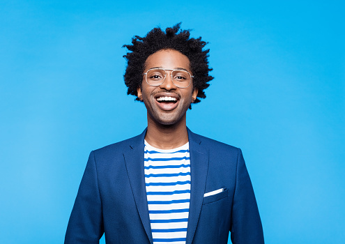 Happy afro american man wearing blue jacket, striped t-shirt and glasses smiling at camera. Studio shot on blue background. Portrait of designer.