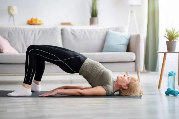 Photo of Stay at home fitness. Strong mature woman doing half bridge yoga pose, strengthening her abs muscles indoors, copy space