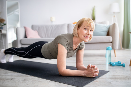 Home training concept. Smiling senior woman doing elbow plank on yoga mat in living room. Positive mature lady doing her workout routine, exercising indoors, strengthening her core muscles