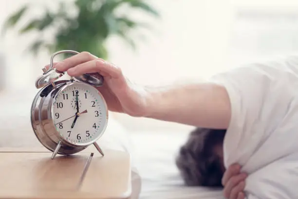 Alarm clock morning wake-up time on bedside table with man reaching to turn off