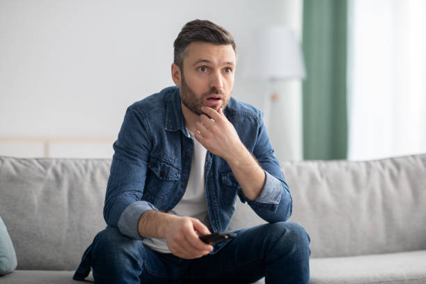 hombre conmocionado con tv remota sentado en el sofá en casa - men one person surprise looking fotografías e imágenes de stock