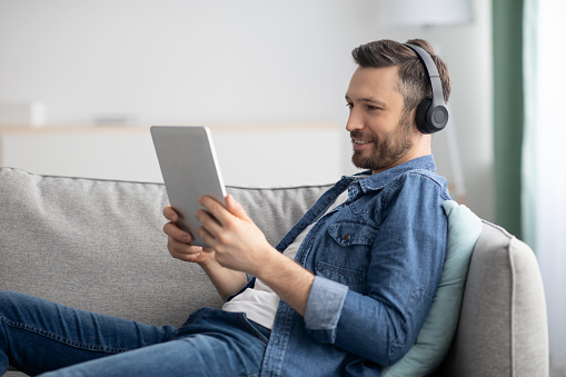 Joyful middle-aged bearded man laying on couch in living room, usind wireless headset and digital tablet, listening to music or watching videos online, using mobile apps, copy space