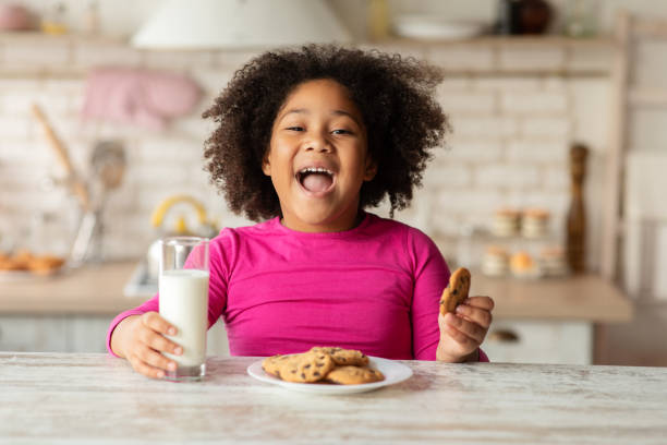 cheerful little black girl sitting at table enjoying milk and cookies - milk child drinking little girls imagens e fotografias de stock