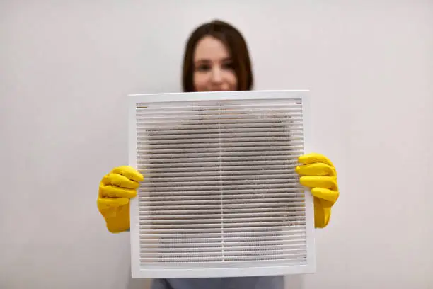Woman holds ventilation grill with dust filter to clean it. Extremely dirty and dusty white plastic, harmful for health. Housewife in protective yellow rubber gloves and blue-grey uniform, blurred.