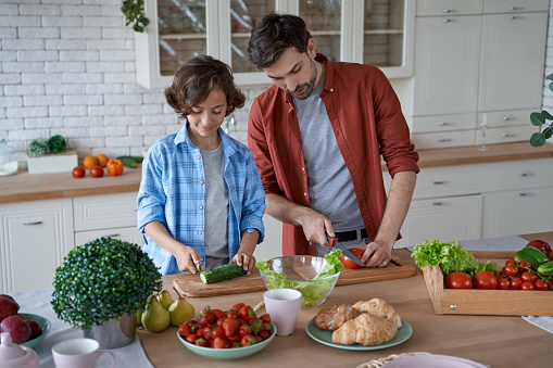 Preparing lunch together. Young father teaching his son how to cut fresh vegetables, preparing salad while standing in the modern kitchen at home. Happy family concept. Cooking with dad