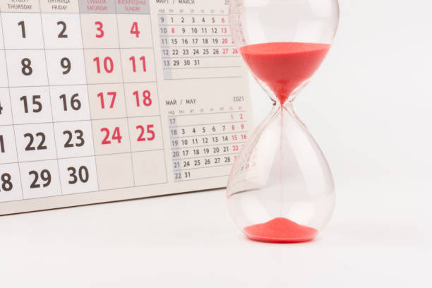 istock Time passing concept. Crystal hourglass with red sand and calendar on light background 1307957860