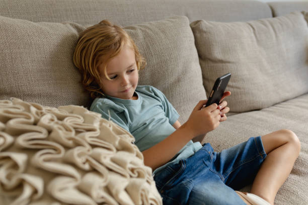 istock Caucasian boy with long blonde hair sitting on couch using smartphone 1307966529