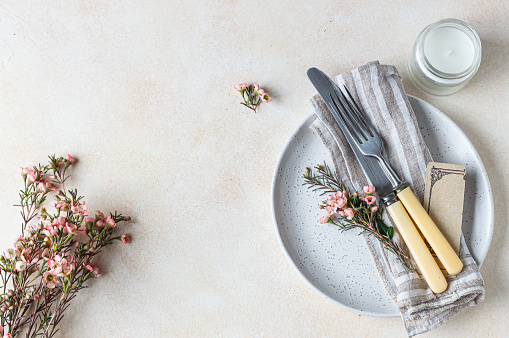 Romantic or spring table setting. Knife and fork, little pink flowers and linen napkin on a plate, light concrete background. Flat lay. Selective focus.
