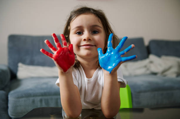 adorable niña mostrando sus manos pintadas con pinturas rojas y azules, sonríe lindamente a la cámara. hobby creativo - oil painting painted image art studio fun fotografías e imágenes de stock