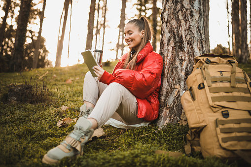 Beautiful woman taking a break, drinking hot tea and using digital tablet while hiking. Success woman hiker hiking on sunrise mountain peak - Young woman with backpack rise to the mountain top. Discovery Travel Destination