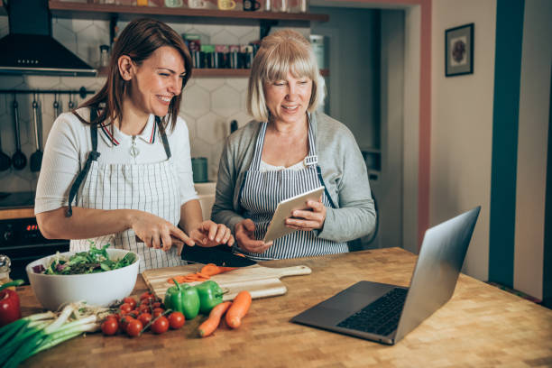 Mother and daughter using laptop while cooking healthy meal in the kitchen.
