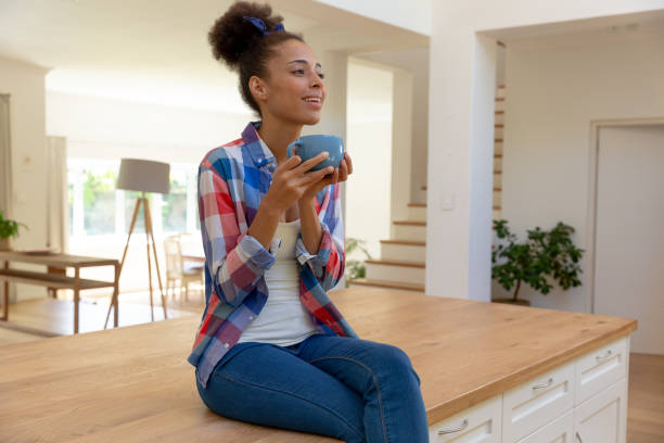 istock Mixed race woman sitting on worktop drinking coffee and smiling in a kitchen 1307992533