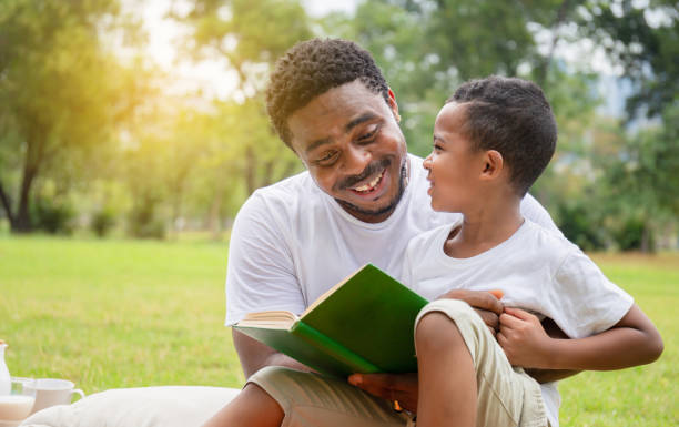 fröhliche afrikanische amerikanische junge und vater bei einem picknick im park, glücklich sohn und vater lesen ein buch, glück familienkonzepte - lesen stock-fotos und bilder