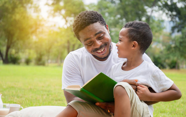 garçon et papa afro-américains gais ayant un pique-nique dans le parc, fils heureux et père affichant un livre, concepts de famille de bonheur - family reading african descent book photos et images de collection