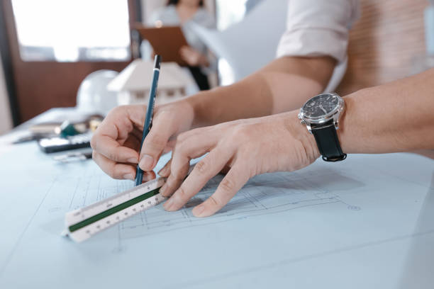 istock engineers holding a pen pointing to a building and  drawing outlay construction plan as guide for builders with details. Engineer design and writing blueprint. 1308042948