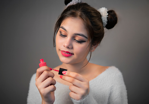 Pretty happy young woman with two hairs bon standing against a dark gray background and applying pink color nail polish against on finger nail.