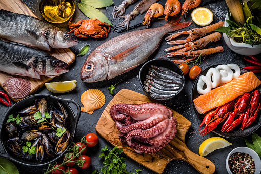 Top view of a multicolored background made of various kinds of fresh fish and seafood such as sea bream, bass, anchovies, salmon, crab, octopus, shrimps, oyster, squid and crayfish and some fresh aromatic herbs. Studio shot taken with Canon EOS 6D Mark II and Canon EF 100 mm f/ 2.8