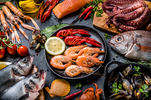 Top view of a multicolored background made of various kinds of fresh fish and seafood such as sea bream, bass, anchovies, salmon, crab, octopus, shrimps, oyster, squid and crayfish and some fresh aromatic herbs. Studio shot taken with Canon EOS 6D Mark II and Canon EF 100 mm f/ 2.8