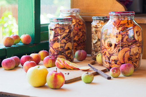 sliced apples on a chopping board and dried apples in jars on a wooden table near the window