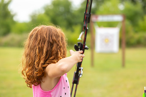 Rear view of a young girl practicing archery. In this image she has just released an arrow, which can be seen flying towards the target. The girl has curly copper colored hair.