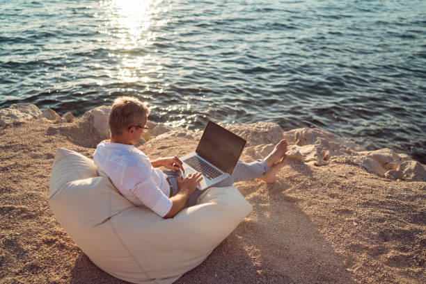 istock Senior man working on his laptop lying on deck chair on the beach 1308090704