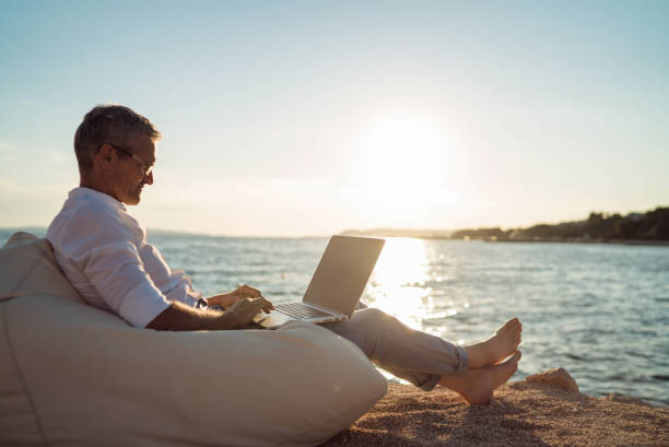 Senior man working on his laptop lying on deck chair on the beach Senior man working on his laptop lying on deck chair on the beach during sunset beach relax stock pictures, royalty-free photos & images