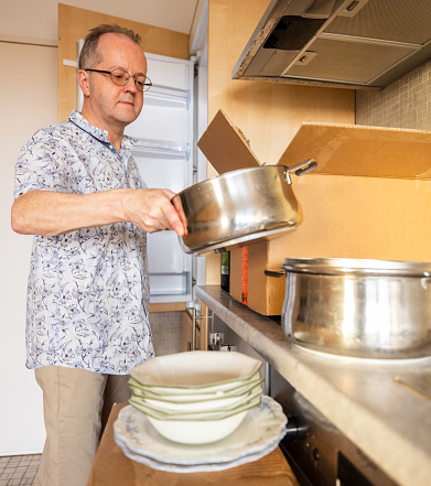 Single senior man moving house in the kitchen while moving in or moving out, unpacking plates and cooking pans from cardboard boxes