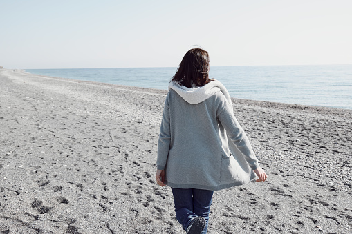 Back view of woman walking on the deserted winter beach, enjoying sunny weather