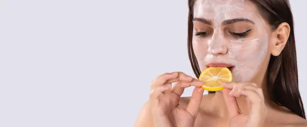 Young brunette with a whitening mask on her face eating an orange slice against a gray banner background. Skin care and vitamins concept. High quality photo