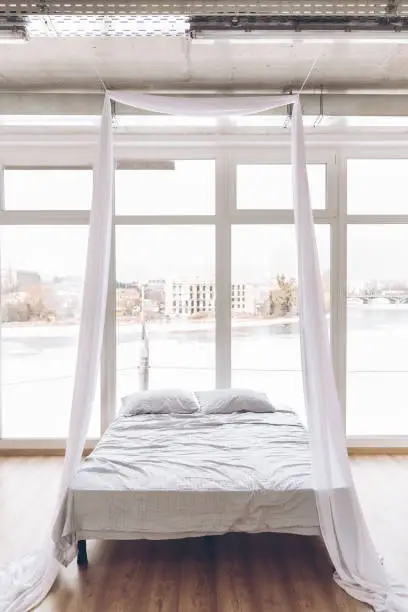 White four-poster bed against the backdrop of a panoramic window with a beautiful view of the city. Modern bedroom interior.