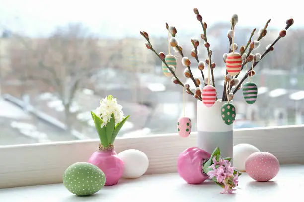 Easter decorations on windowsill in Spring. Wooden painted eggs hang on pussy willow twigs in ceramic vase. Easter eggs in pastel colors, pink and green. Urban view over town, cityscape in window.