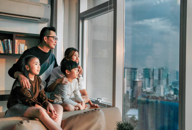 asiatische chinesische familie mit 2 kindern, die durch das fenster schauen und den wunderschönen sonnenuntergang im wohnzimmer in kuala lumpur stadt genießen - skyscraper city life urban scene building exterior stock-fotos und bilder