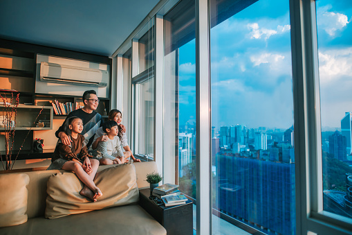 asian chinese family with 2 children looking through window enjoying beautiful sunset at living room in kuala lumpur city