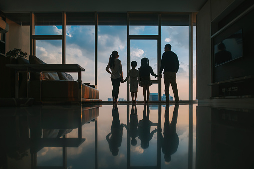 asian chinese family with 2 children looking through window enjoying beautiful sunset at living room in kuala lumpur city