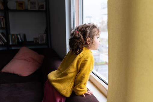 Portrait of cute 2,5 years old girl wearing a yellow sweater looking through window. Shot indoor with a full frame mirrorless camera.