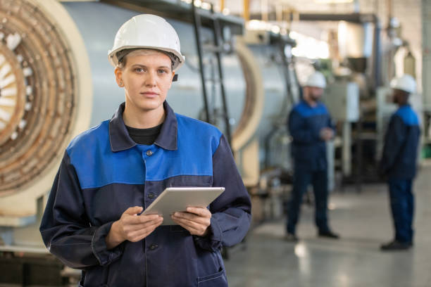 istock Young female factory worker in overalls and hardhat using tablet in working environment 1308119904