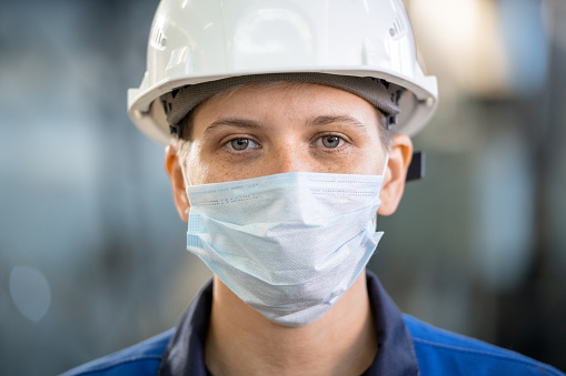 Young female factory worker in overalls, hardhat and protective mask looking at you while standing in workshop in front of camera