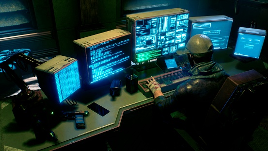 A male hacker surrounded by glowing monitors hacks into someone else's computer network in a dark room of his office. A view of the hacker's futuristic computer office.