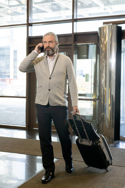 istock Mature businessman with smartphone by ear pulling suitcase while entering hall of hotel 1308125685
